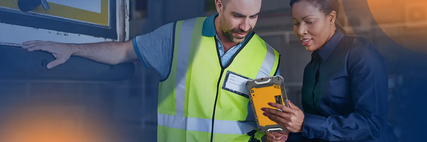 Two workers in a warehouse setting review information together on a tablet near a loading dock door.